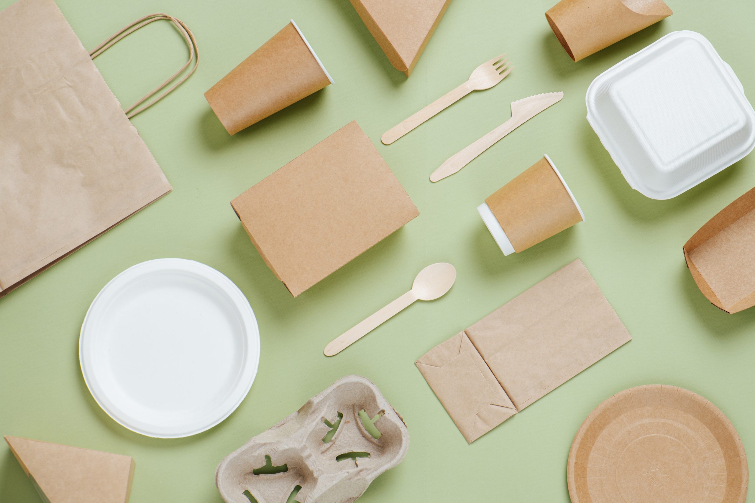 Eco-friendly disposable utensils made of bamboo wood and paper on a green background. Draped spoons, fork, knives, bamboo bowls with paper cups and packet
