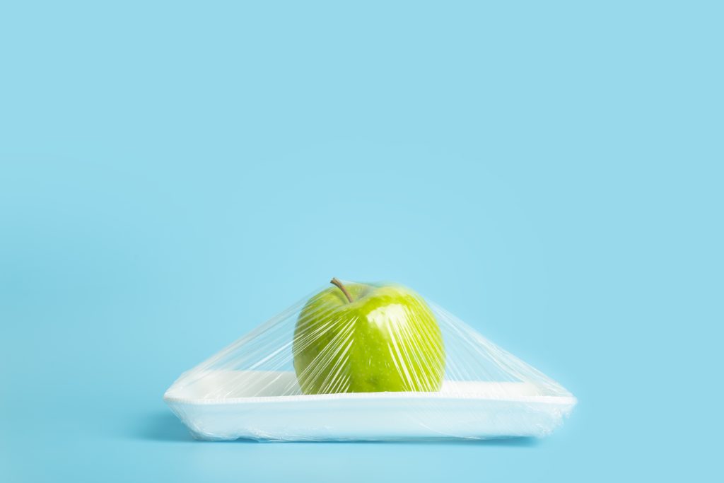 Fruits in plastic packaging from the supermarket are minimal. Apple in cellophane and non-degradable plastic on a blue background. Biodegradable product packaging, environmental protection, nature pollution. High quality photo
